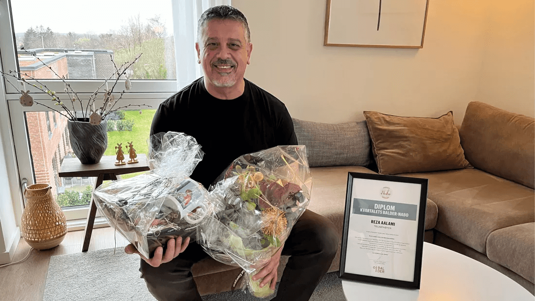 Reza, a resident in Triumphbyen, sits in his living room with flowers and a certificate.