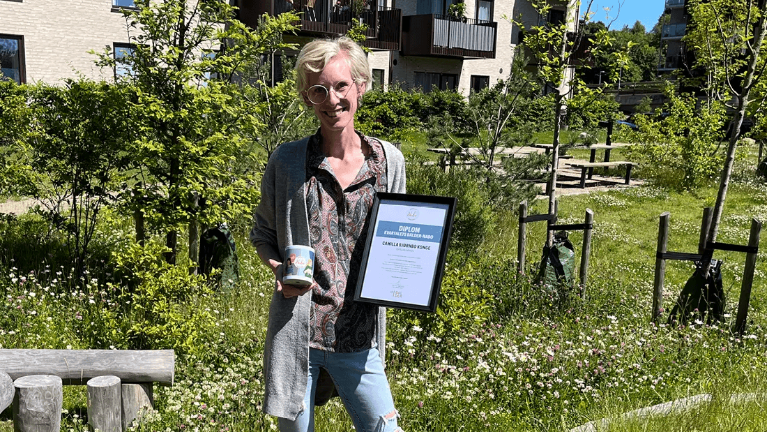 Camilla, a resident at Ofeliahaven, stands in the courtyard with a certificate and a cup of coffee.