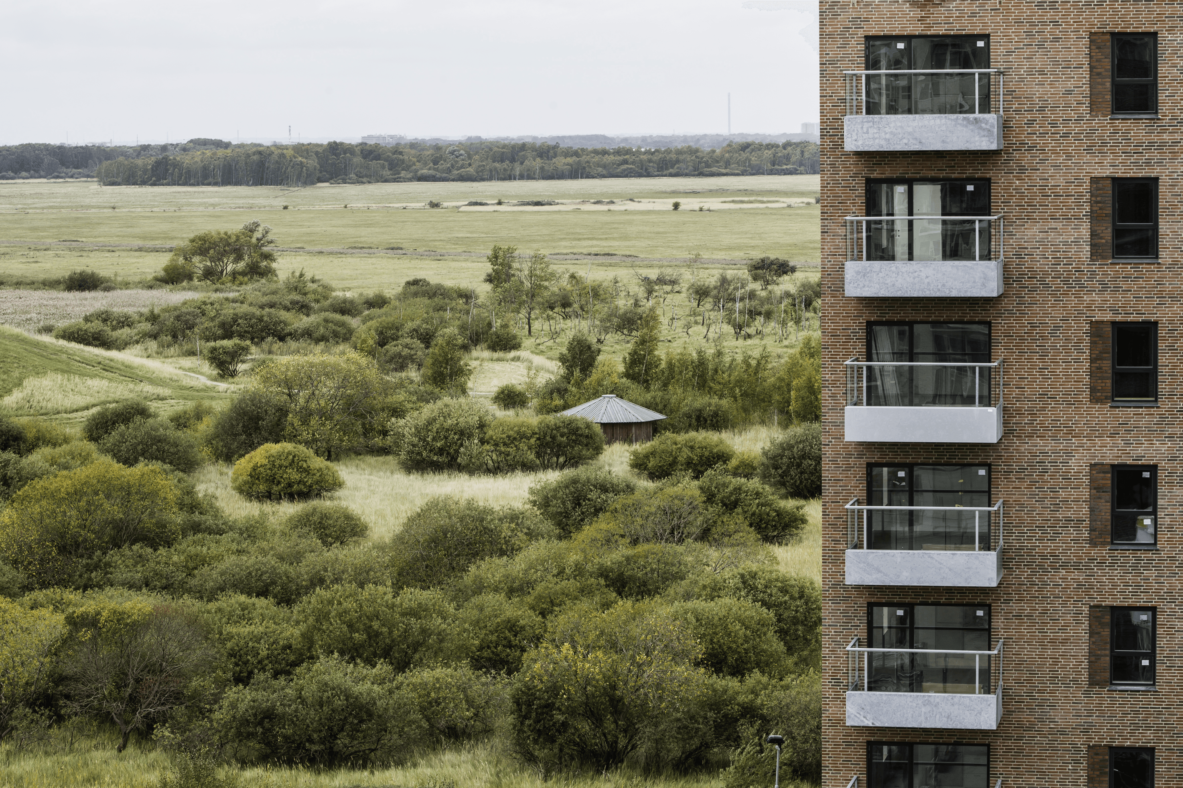 Elsehus exterior photo showing a brick building façade with balconies on the right and a wide view over green nature with shrubs, trees, and open fields.